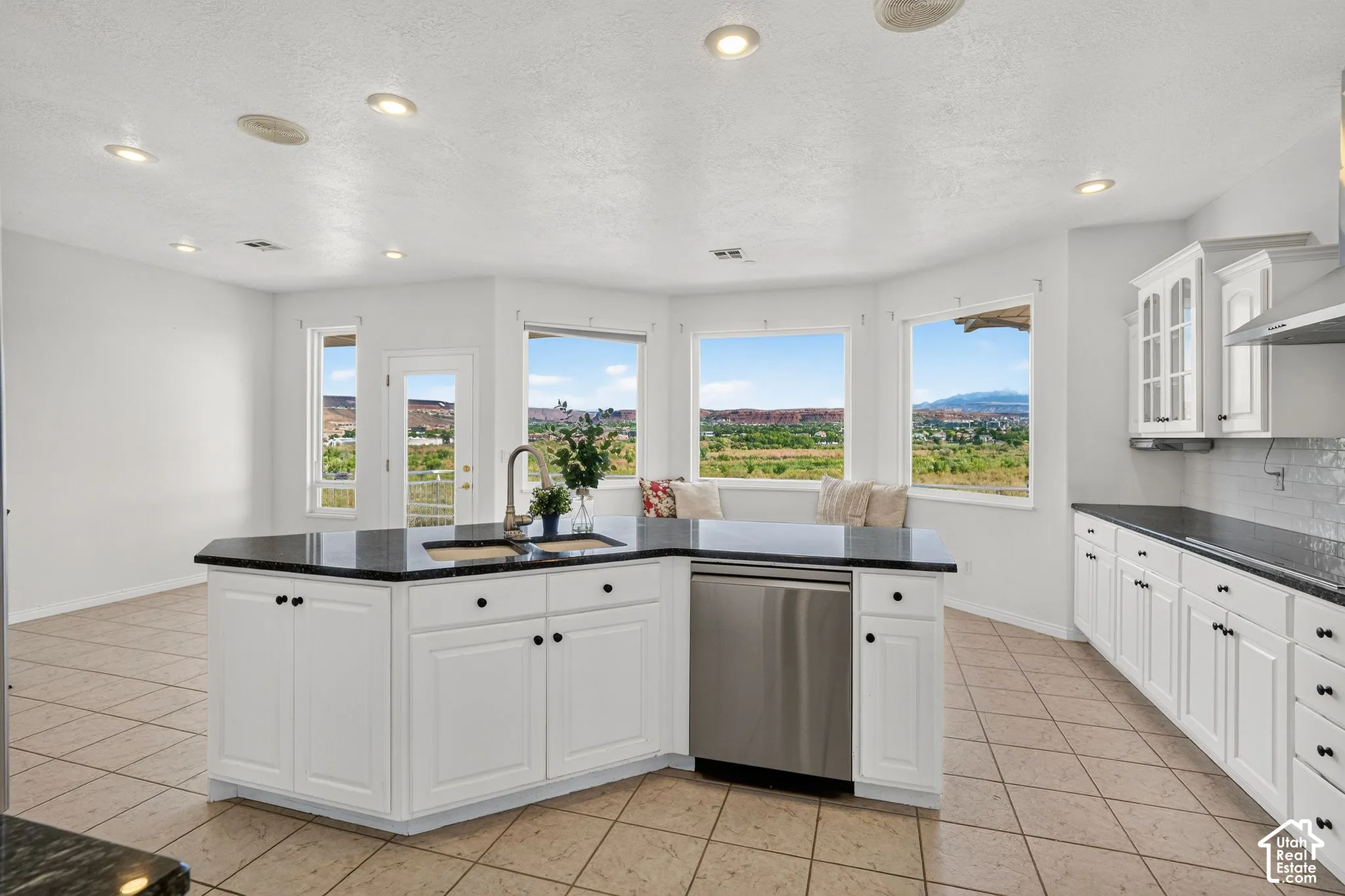 Bright, sun-filled kitchen featuring white shaker cabinetry with black hardware, a curved island with black countertop and stainless steel dishwasher, recessed ceiling lighting, and expansive multi-pane windows framing scenic landscape vistas