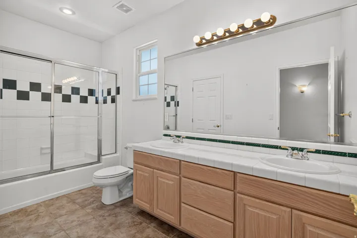 Dual vanity with white tile countertop, extensive wall mirror, and integrated lighting fixture