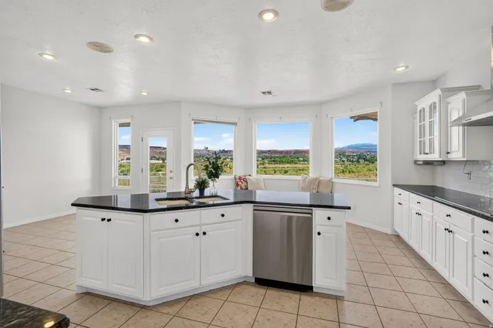 Bright, sun-filled kitchen featuring white shaker cabinetry with black hardware, a curved island with black countertop and stainless steel dishwasher, recessed ceiling lighting, and expansive multi-pane windows framing scenic landscape vistas