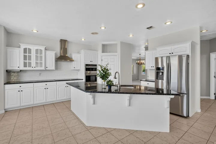 Chef's kitchen featuring white cabinetry with glass-front uppers, stainless steel appliances including double wall ovens and a professional-style range hood, quartz countertops, a large center island with seating, recessed ceiling lighting, and tile floor