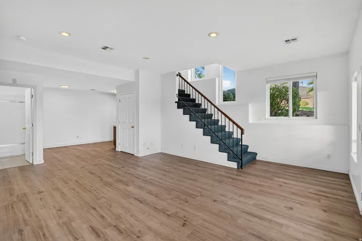 Expansive room featuring wood-finish flooring, recessed lighting, a carpeted staircase with an iron railing, and multiple windows
