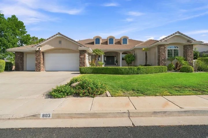 Elegant single-story residence featuring a stucco and brick exterior with multiple roof dormers, two-car garage, and manicured front landscaping with mature trees and hedging
