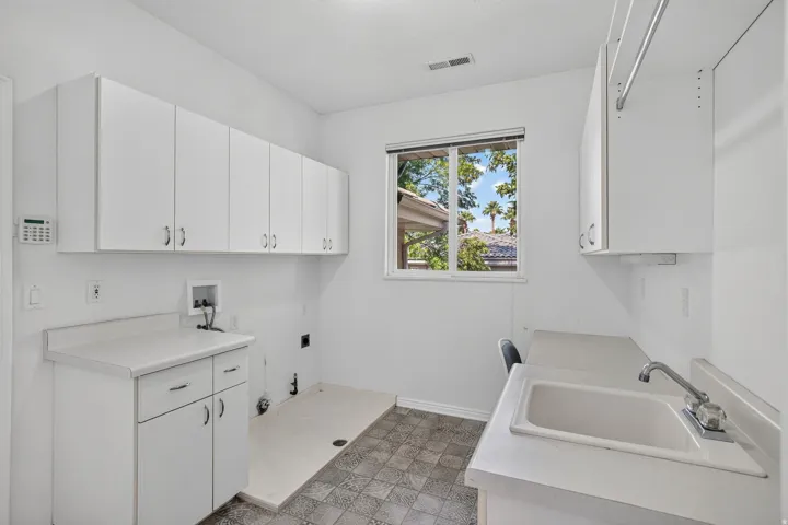 Bright laundry room featuring expansive white cabinetry with ample storage, dual stainless steel sink basins with chrome faucetry, patterned tile flooring, and abundant natural light from a large picture window overlooking outdoor landscaping