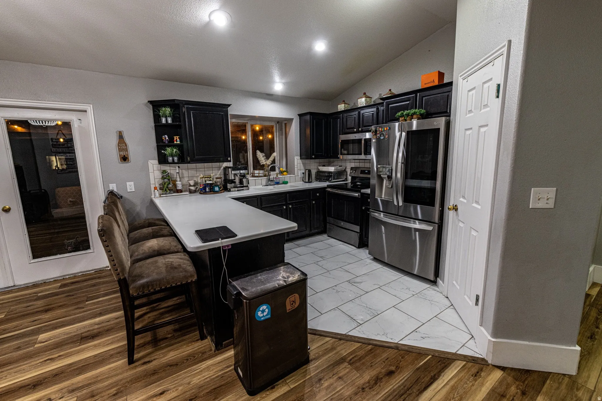 Kitchen featuring black cabinetry, stainless steel appliances, a white tile backsplash, and a breakfast bar with a light-toned countertop