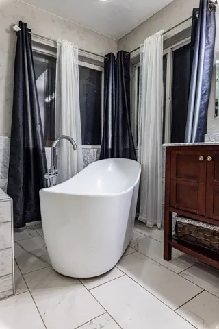 Freestanding soaking tub with floor-mounted faucet, white rectangular tile flooring, and a dark wood vanity with a light-colored countertop