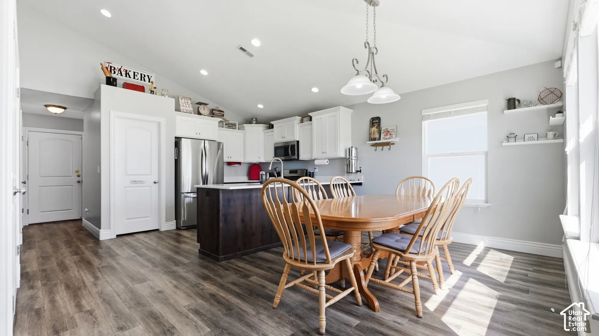 Open-concept kitchen and dining area featuring vaulted ceilings, wood-finish flooring, white cabinetry, and a large window with a deep sill