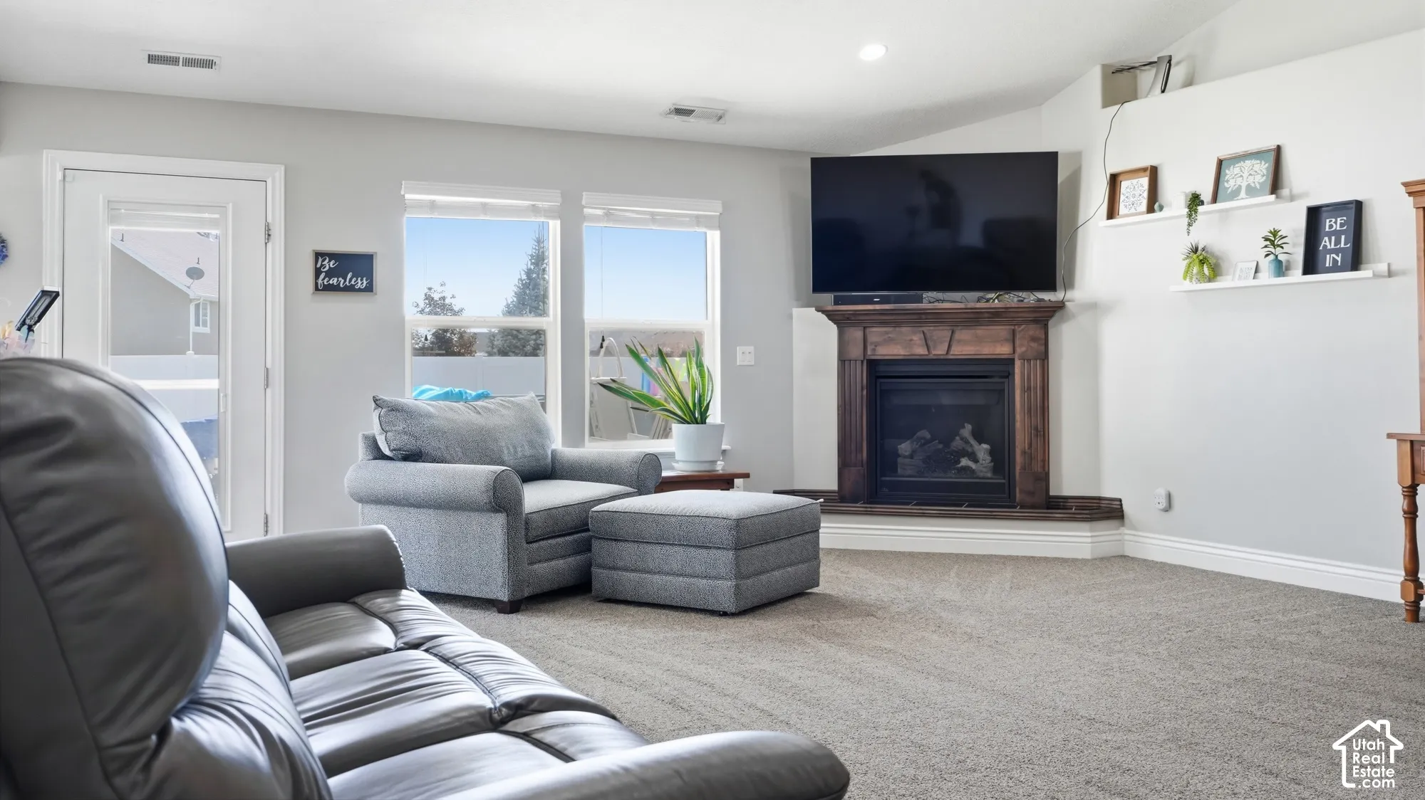 Vaulted ceiling, corner fireplace with wood mantle, carpeted flooring, large windows, and a glass-paneled door
