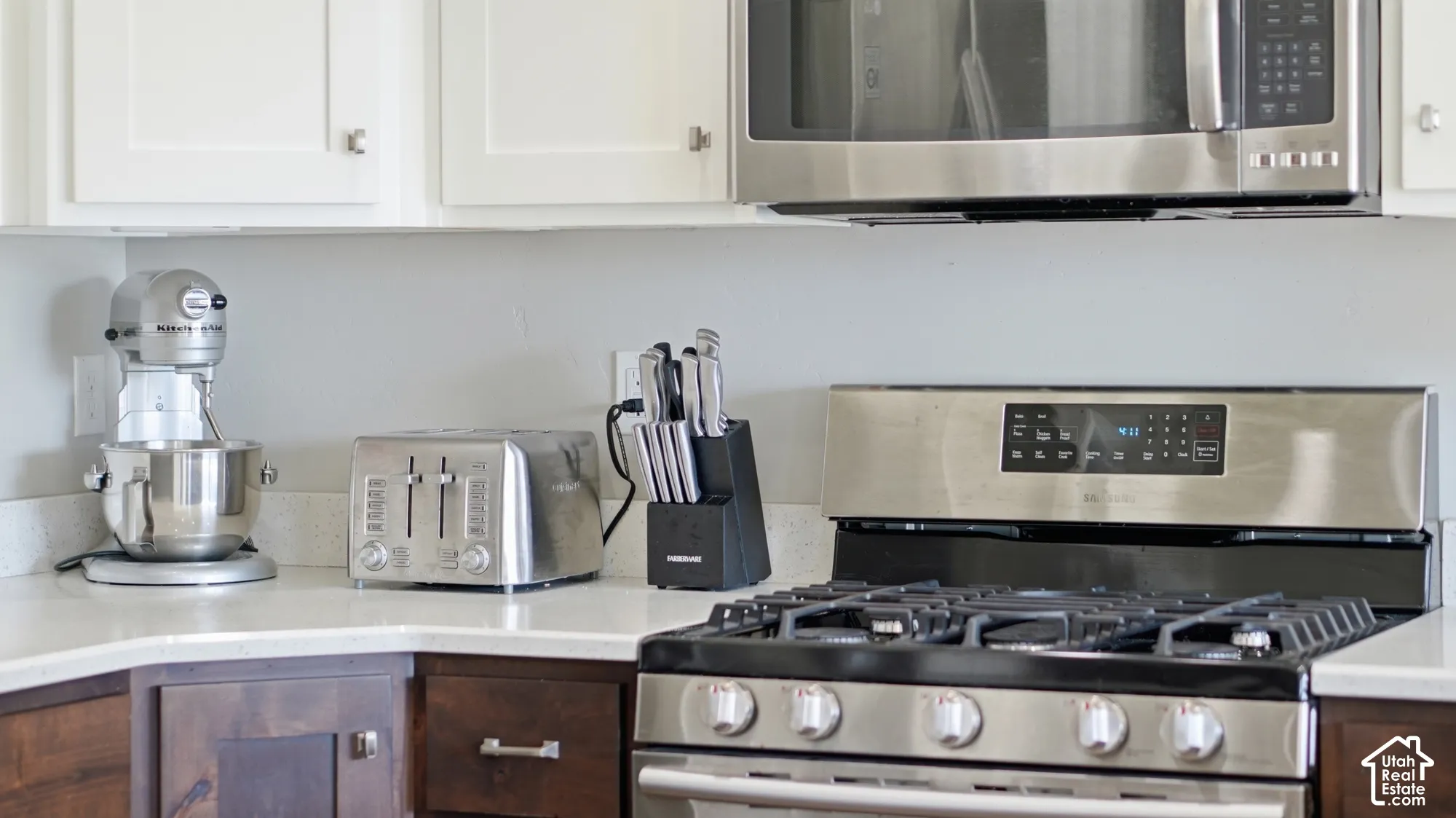 Stainless steel gas range with grates, stainless steel built-in microwave, white upper cabinetry, wood-finish lower cabinetry, and light-toned countertop