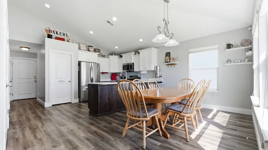 Open-concept kitchen and dining area featuring vaulted ceilings, wood-finish flooring, white cabinetry, and a large window with a deep sill