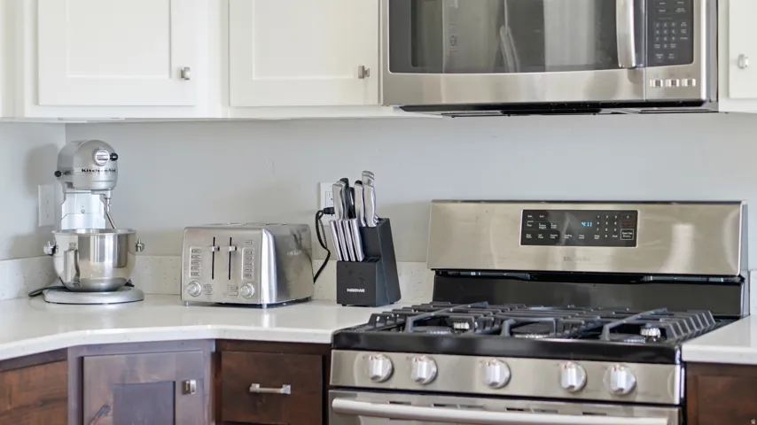 Stainless steel gas range with grates, stainless steel built-in microwave, white upper cabinetry, wood-finish lower cabinetry, and light-toned countertop
