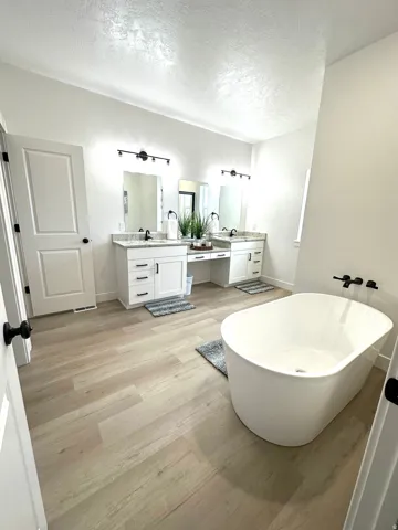 Freestanding oval soaking tub, dual vanity with white cabinetry, granite-finish countertops, black hardware, and wood-finish flooring