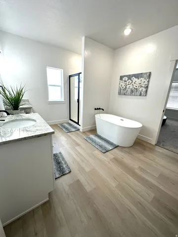 Freestanding soaking tub, walk-in shower with black-framed glass, wood-finish flooring, vanity with integrated sink and stone-look countertop