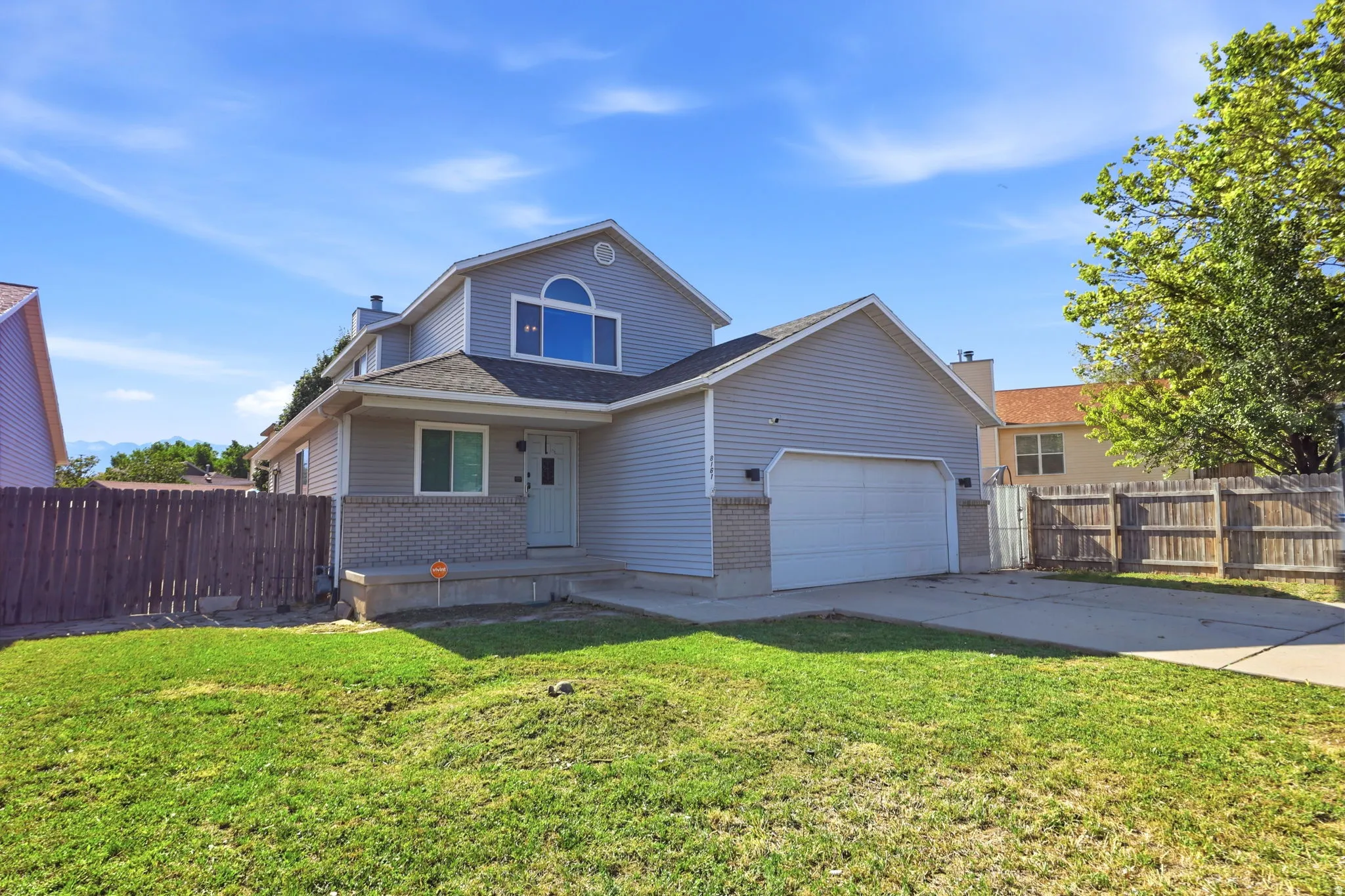 Two-story residence featuring light gray siding, an integrated two-car garage, an arched window on the upper level, a covered front porch, and a wooden privacy fence