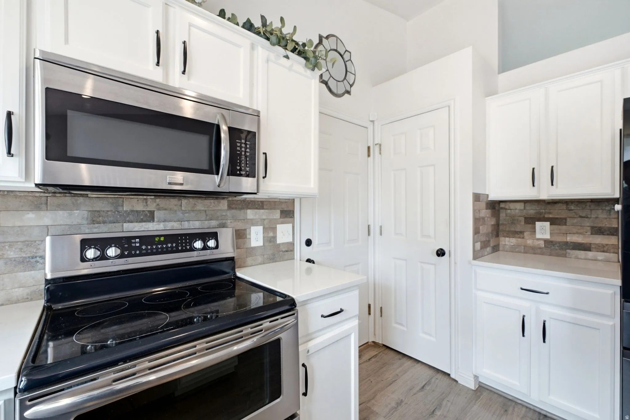 White cabinetry with black hardware, stainless steel appliances, stacked stone backsplash, light-toned countertops, and wood-finish flooring