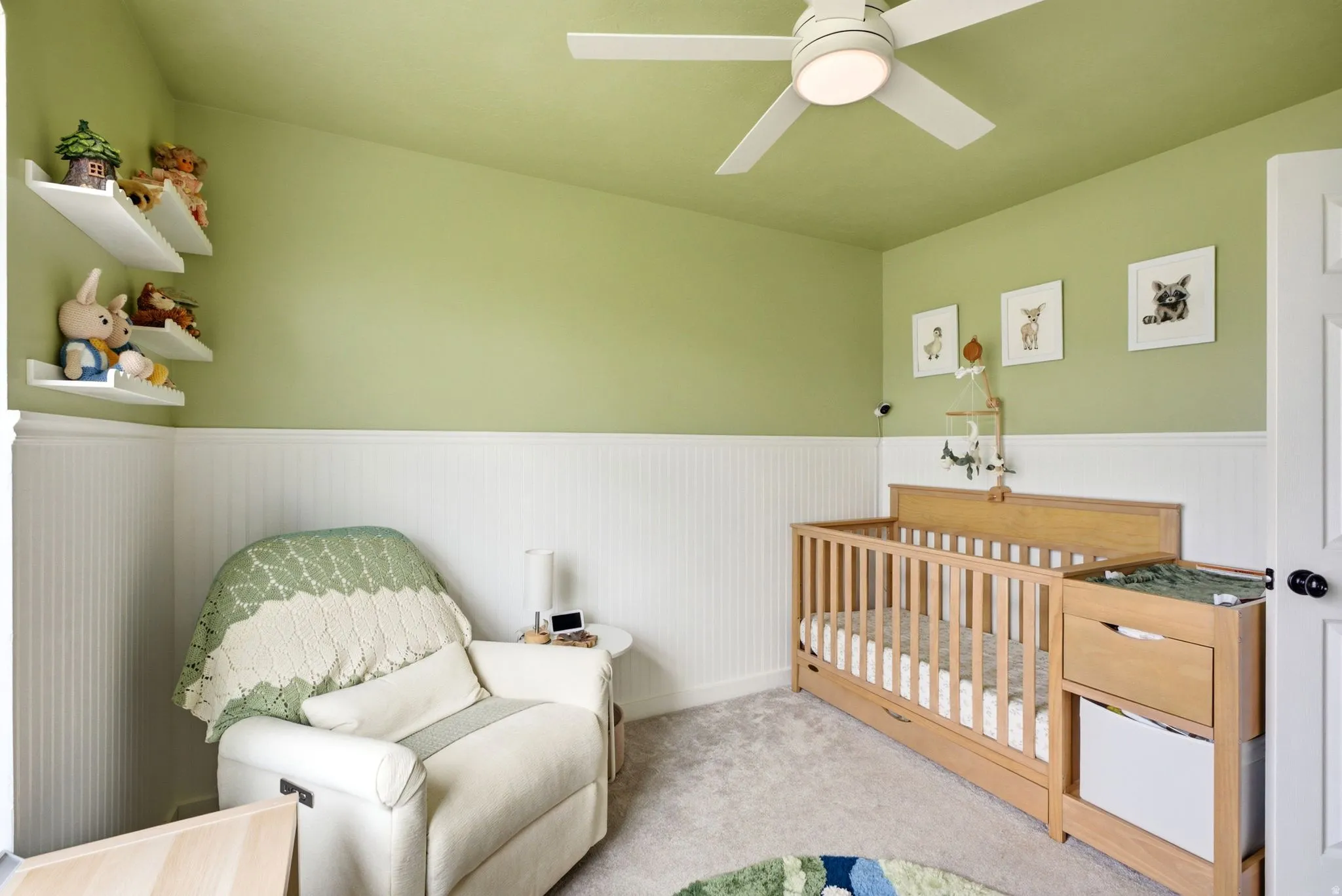 Wall-mounted shelving, beadboard wainscoting, ceiling fan with integrated lighting, wood-finish crib, and light-toned carpeting