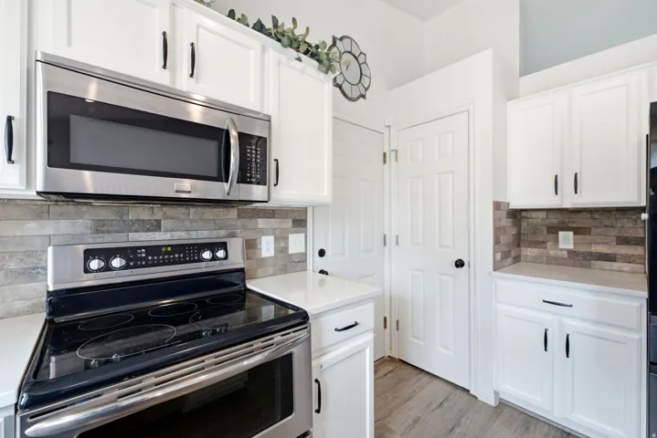 White cabinetry with black hardware, stainless steel appliances, stacked stone backsplash, light-toned countertops, and wood-finish flooring