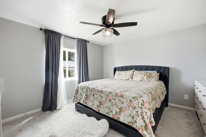 Plush carpet flooring, a window with opaque and sheer curtains, and a ceiling fan with integrated lighting