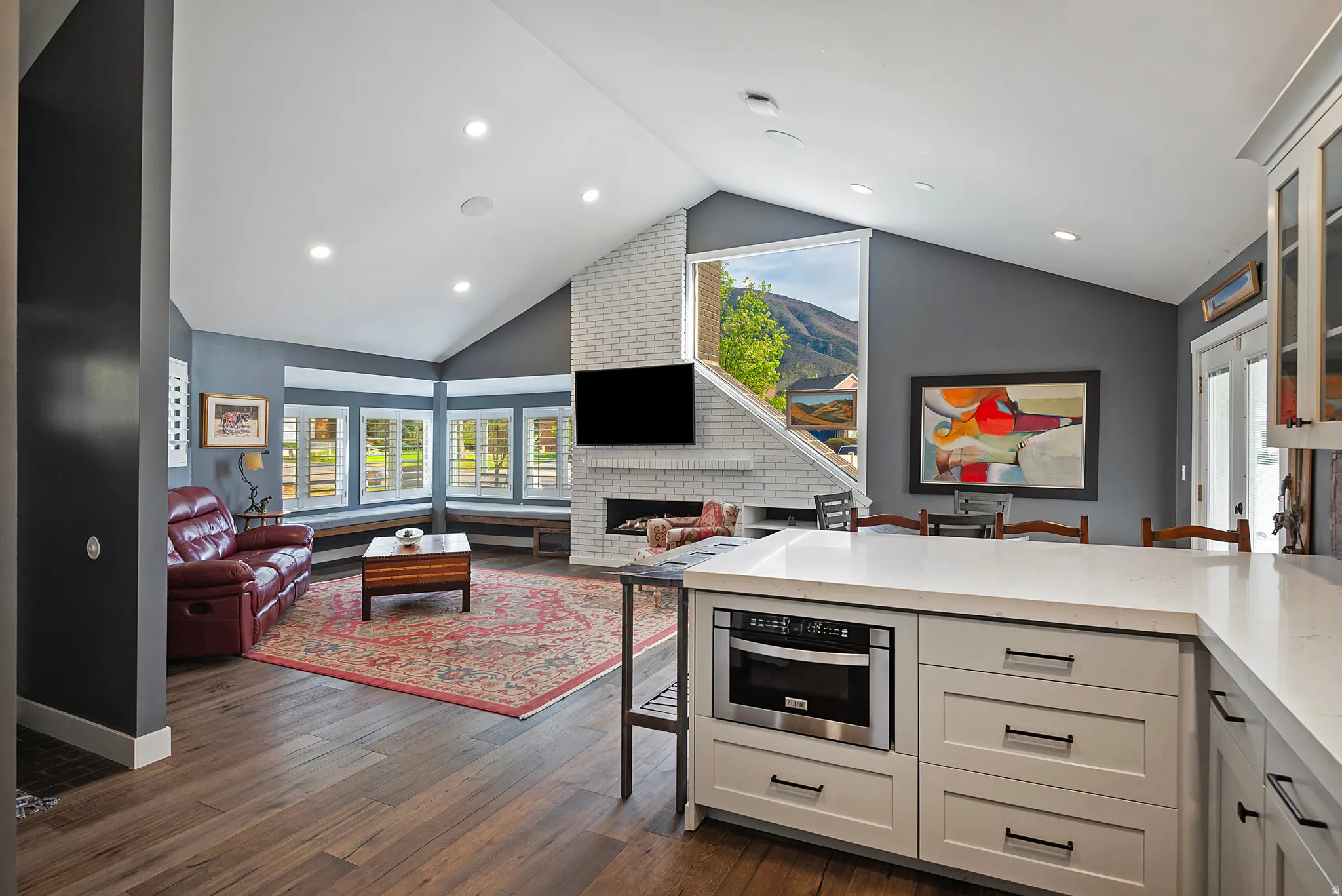 Open-concept kitchen and living area featuring custom cabinetry with integrated appliances, wood-finish flooring throughout, recessed ceiling lighting, and a fireplace with brick surround
