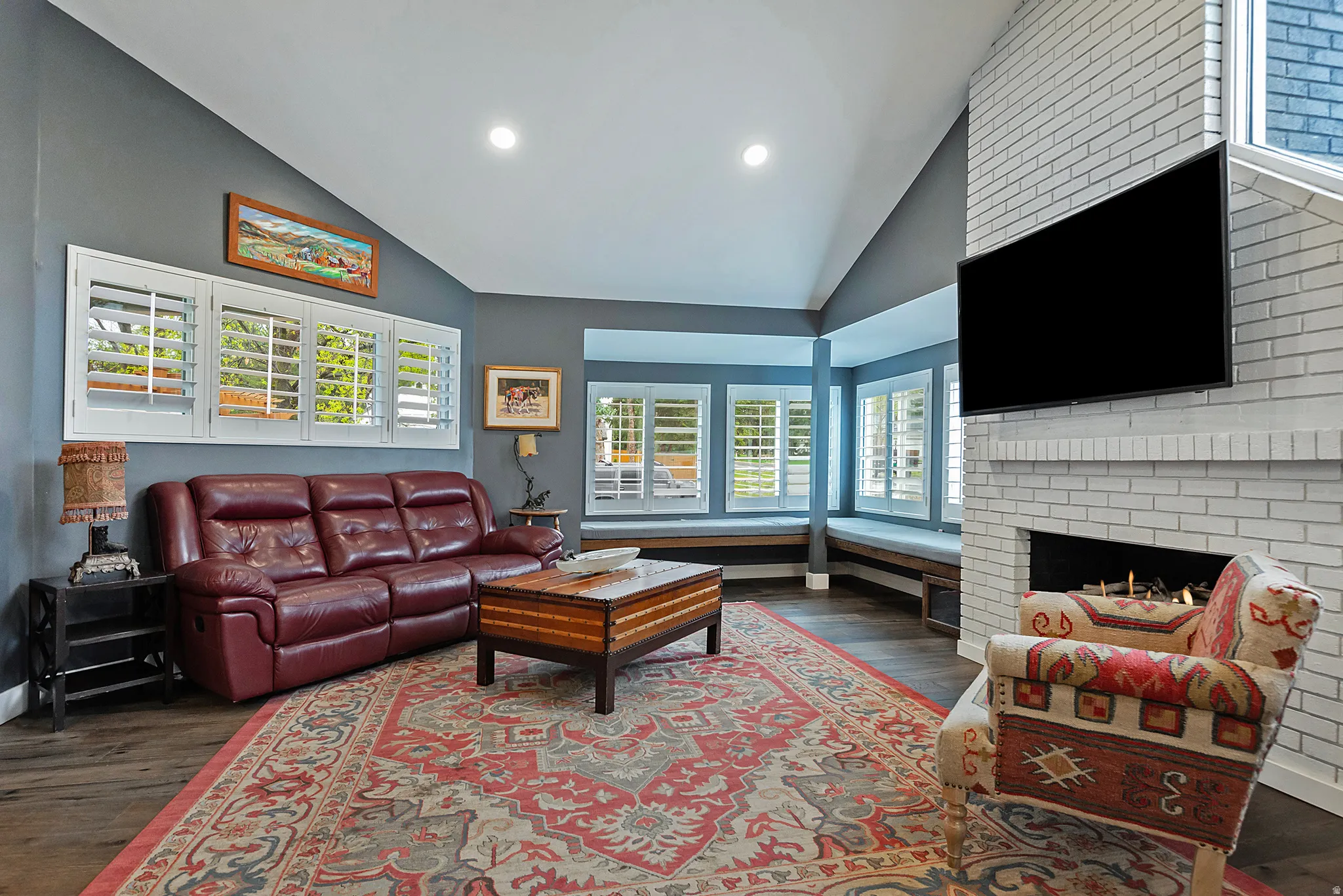 Vaulted ceiling with recessed lighting, wood-finish flooring, white brick fireplace, built-in window seating, and plantation shutters