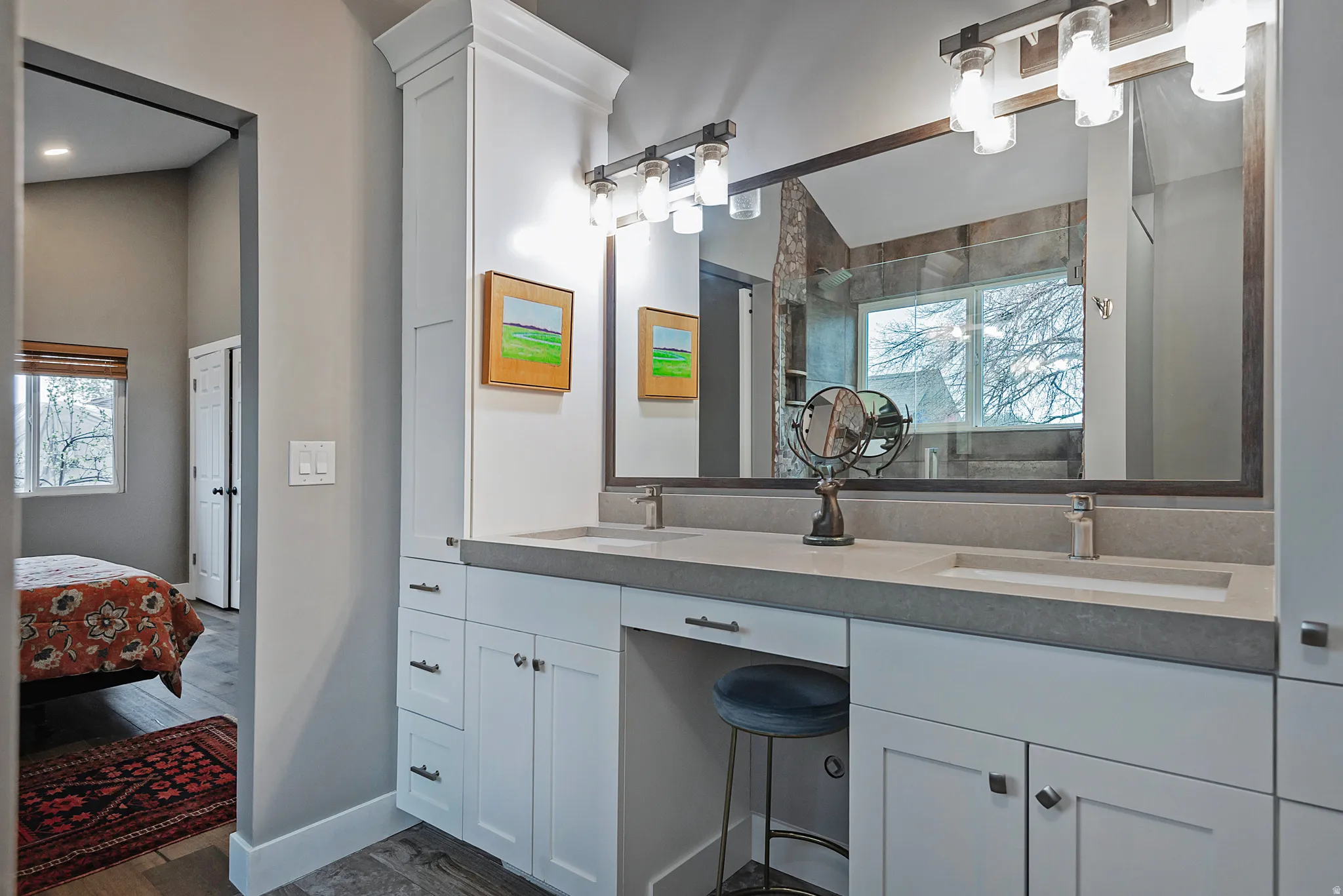 Dual integrated sinks set into a gray countertop, extensive white cabinetry with varied storage, vanity area, and a large wall-mounted mirror with overhead lighting