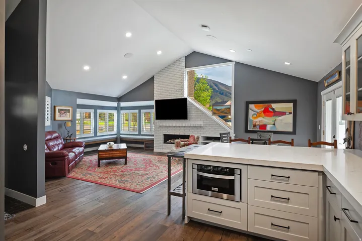 Open-concept kitchen and living area featuring custom cabinetry with integrated appliances, wood-finish flooring throughout, recessed ceiling lighting, and a fireplace with brick surround