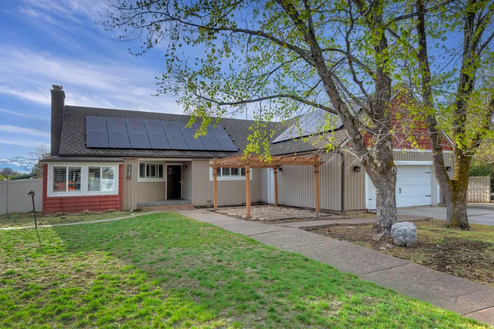 Exterior featuring solar panels, a pergola, an attached garage, a paved walkway, and mature trees