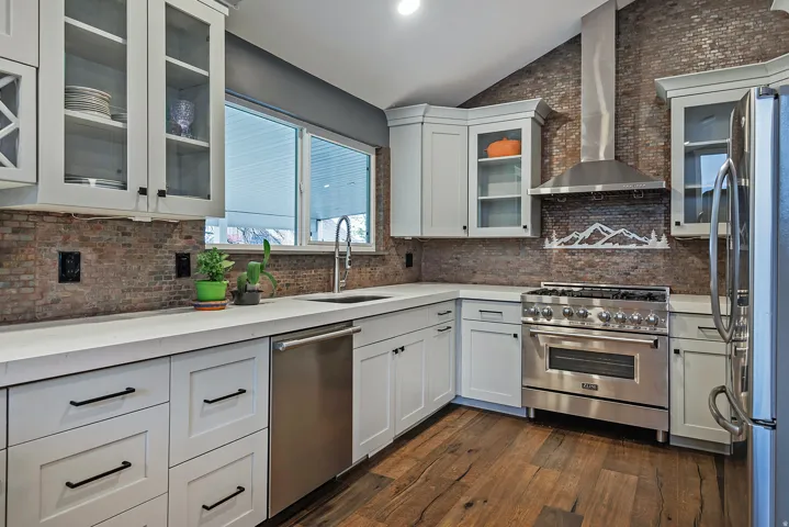 White cabinetry with dark hardware, stone-finish backsplash, stainless steel appliances, wood-finish flooring, and light-toned countertops