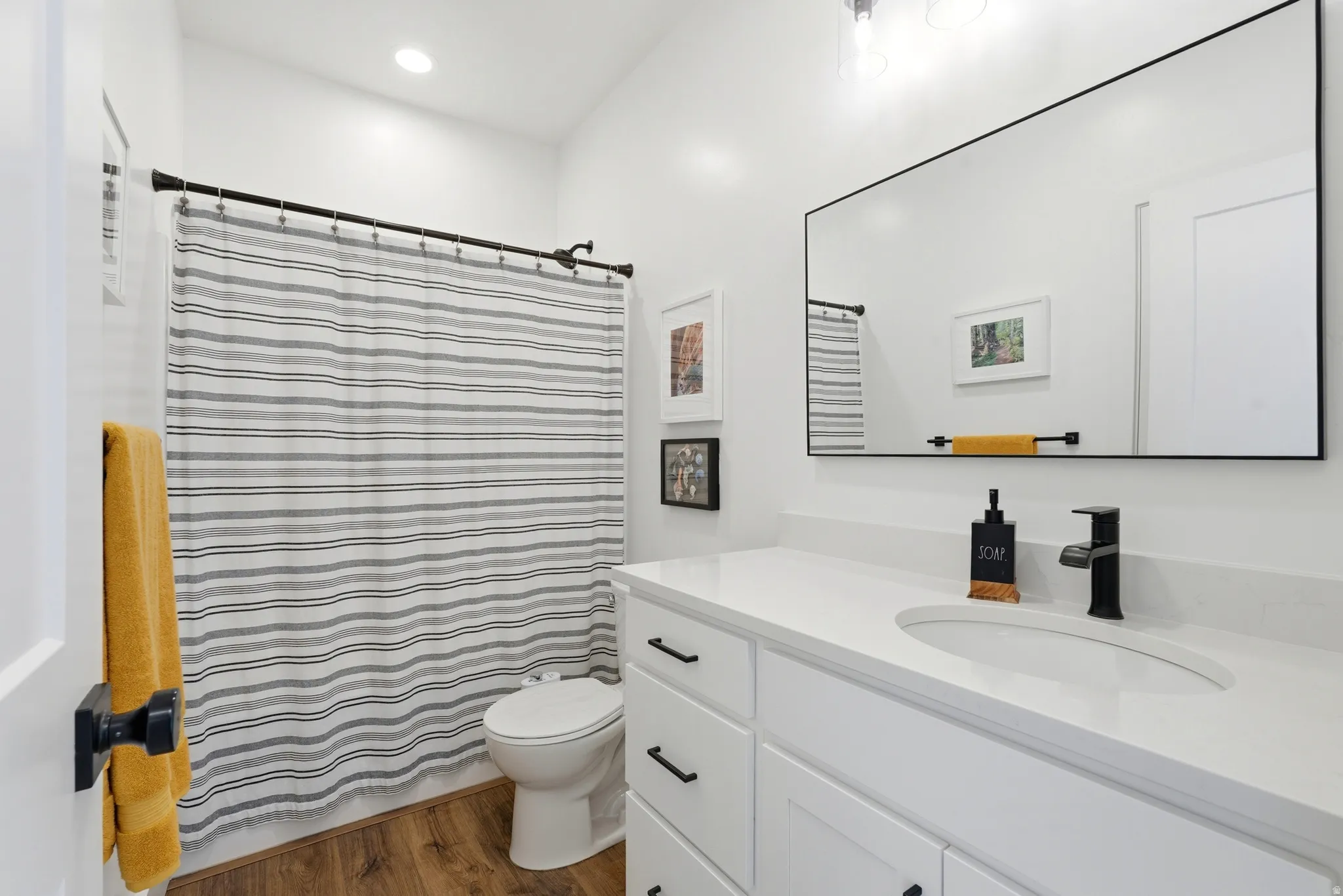 Recessed lighting illuminates a bathroom featuring wood-finish flooring, a white vanity with an integrated sink, a matte black faucet, and a large rectangular mirror with a black frame