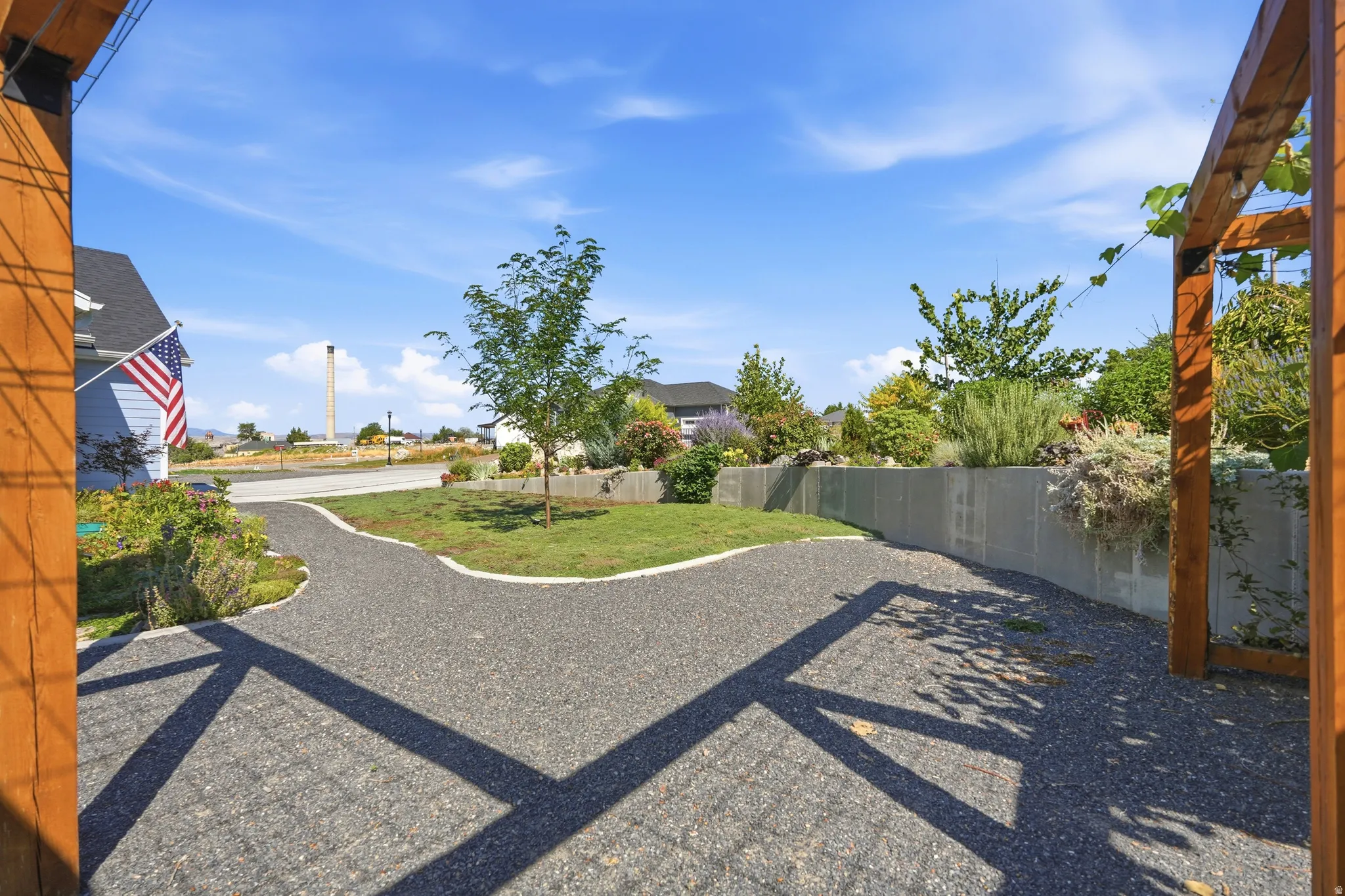 Gravel pathway leading to a grass lawn, featuring a concrete retaining wall with planted shrubs and a wooden pergola structure