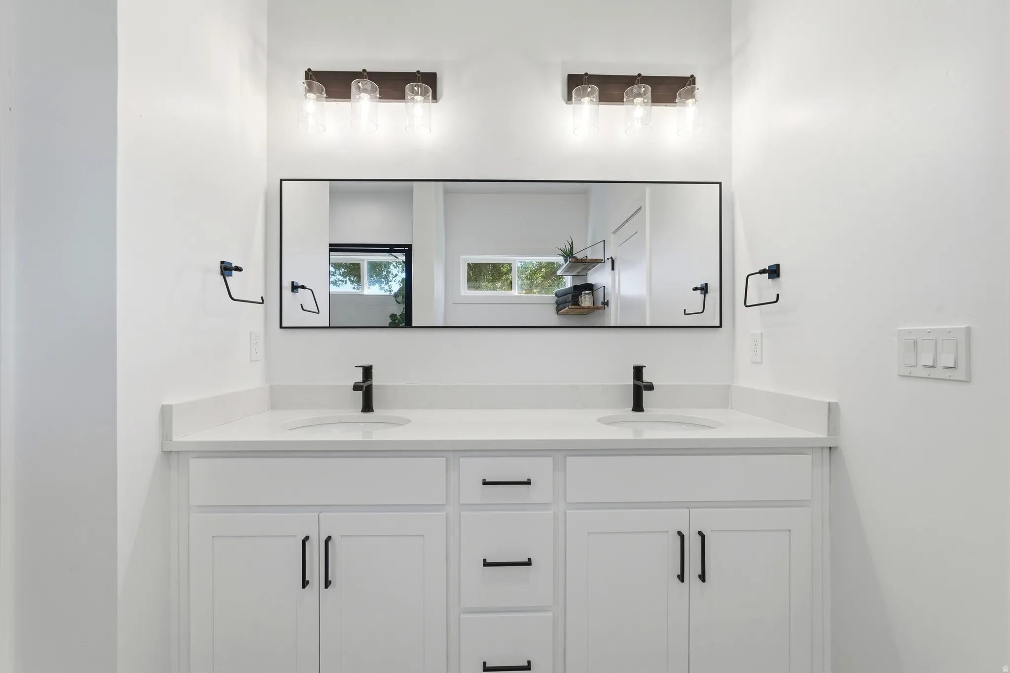 Dual-vanity bathroom featuring a wide quartz countertop, white cabinetry with ample storage, matte black faucets and hardware, and a large framed mirror with integrated lighting