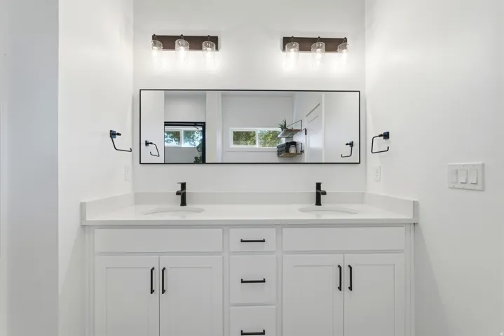 Dual-vanity bathroom featuring a wide quartz countertop, white cabinetry with ample storage, matte black faucets and hardware, and a large framed mirror with integrated lighting