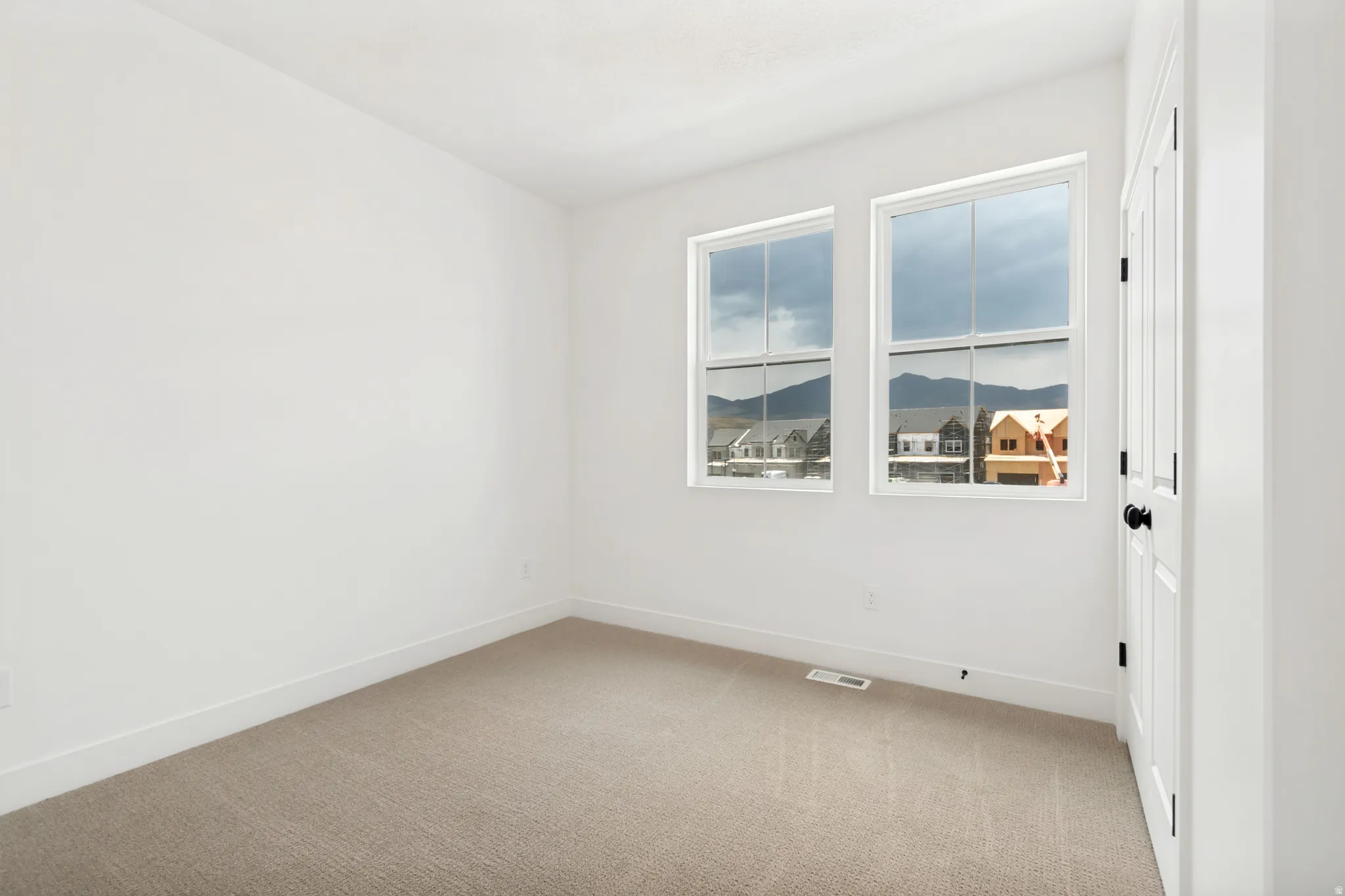 Neutral carpeting throughout, white painted walls, two large windows, mountain scenery, and a white paneled door with a black knob