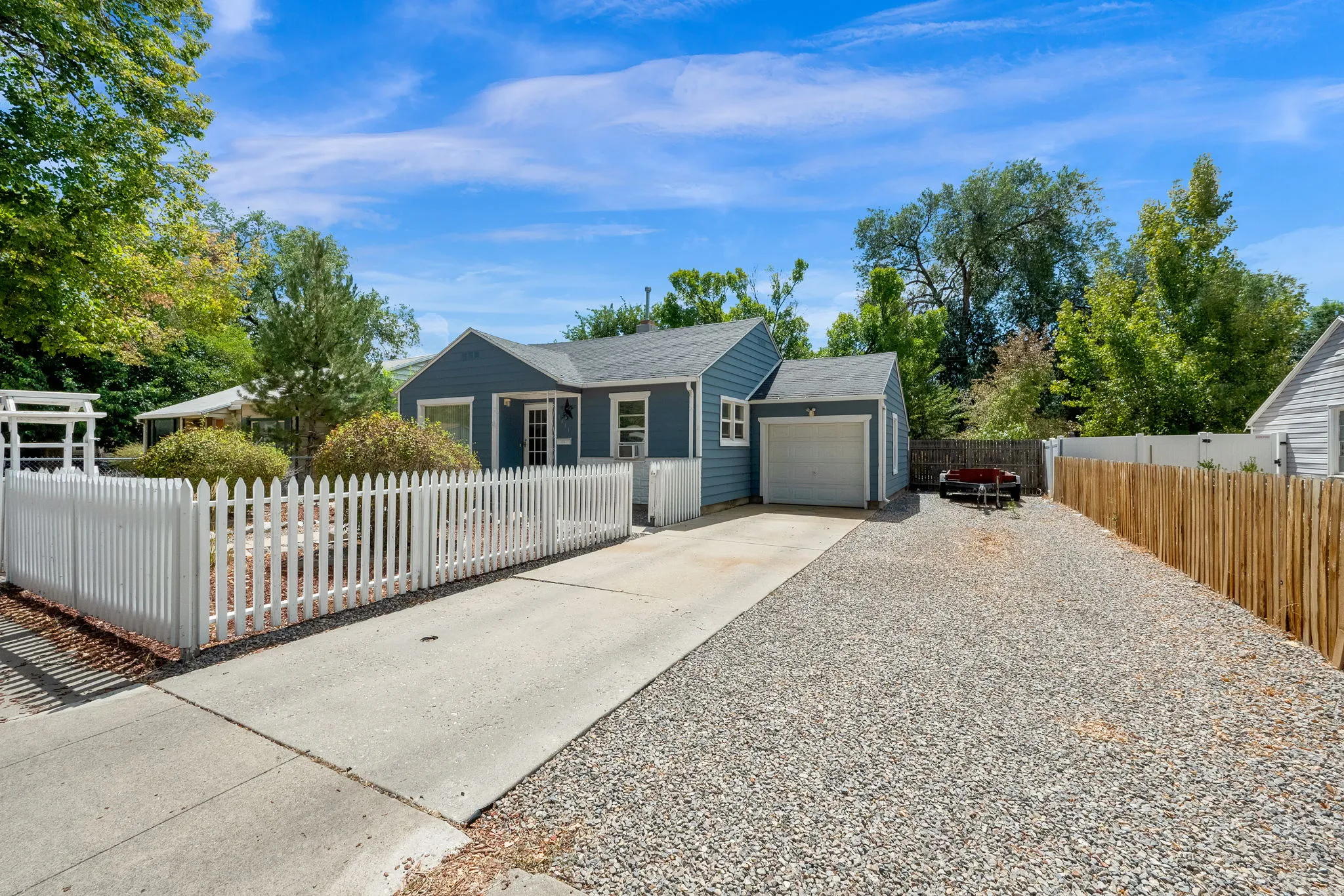 Blue siding exterior with white trim, single-car garage, white picket fence, concrete driveway, and gravel parking area
