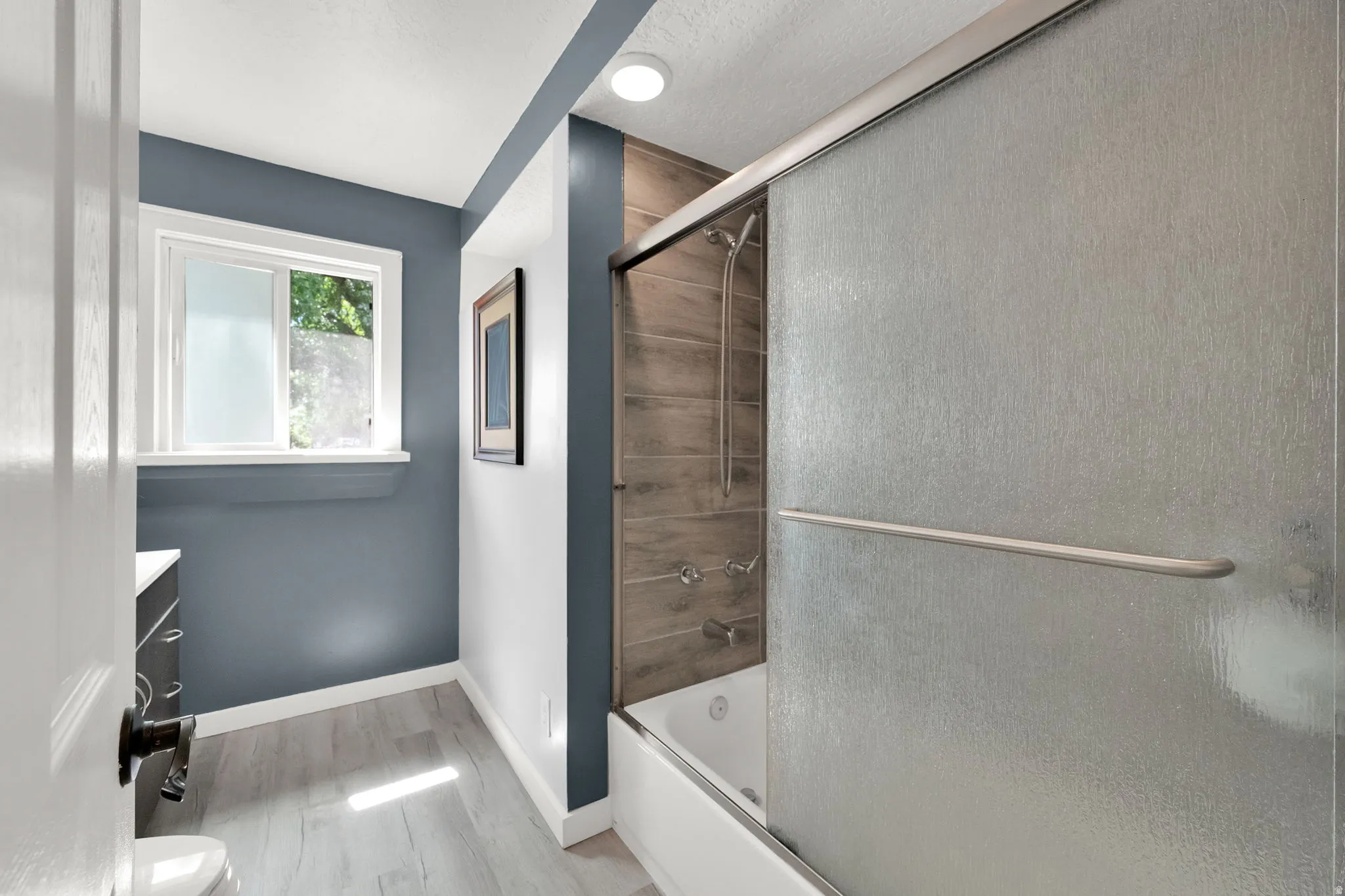 Bathroom featuring wood-finish flooring, a window, a white vanity with a dark countertop, and a shower-tub combination with a sliding glass enclosure and wood-look tile surround