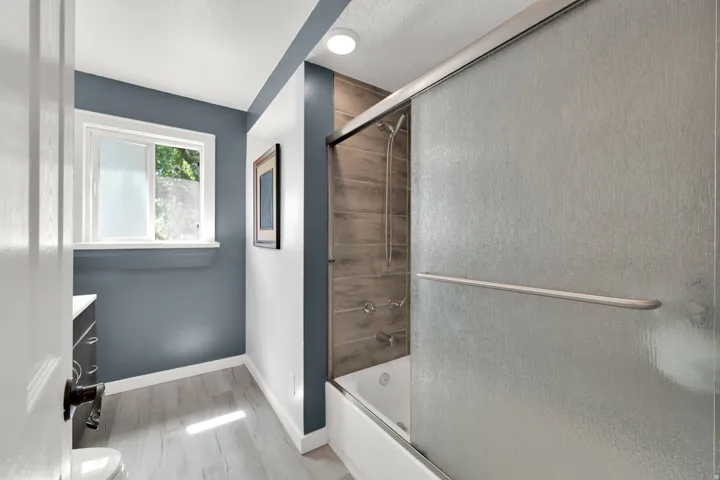 Bathroom featuring wood-finish flooring, a window, a white vanity with a dark countertop, and a shower-tub combination with a sliding glass enclosure and wood-look tile surround