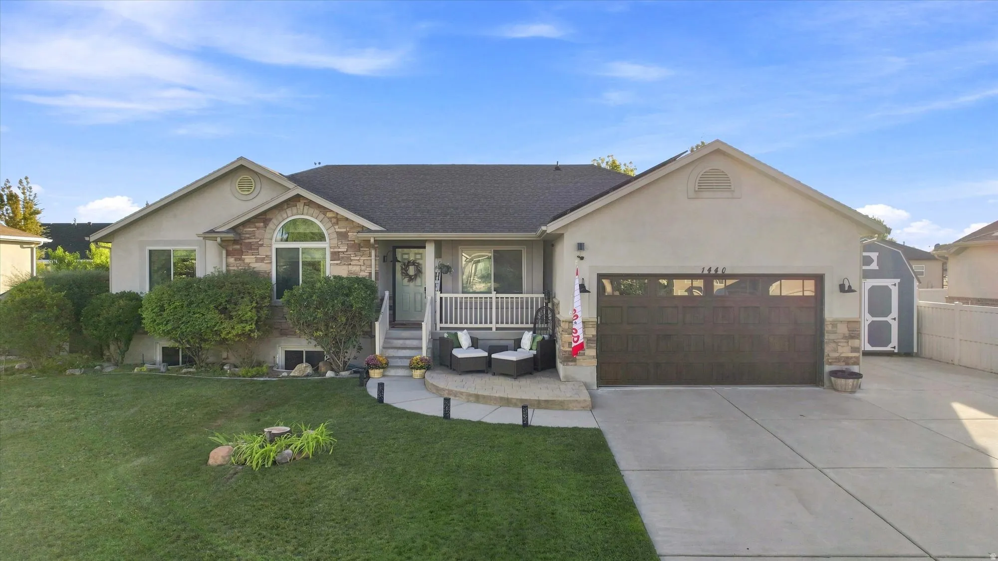 Stone veneer accents on the facade, arched window, attached garage with a dark wood-finish door, covered front porch, and a dedicated storage shed
