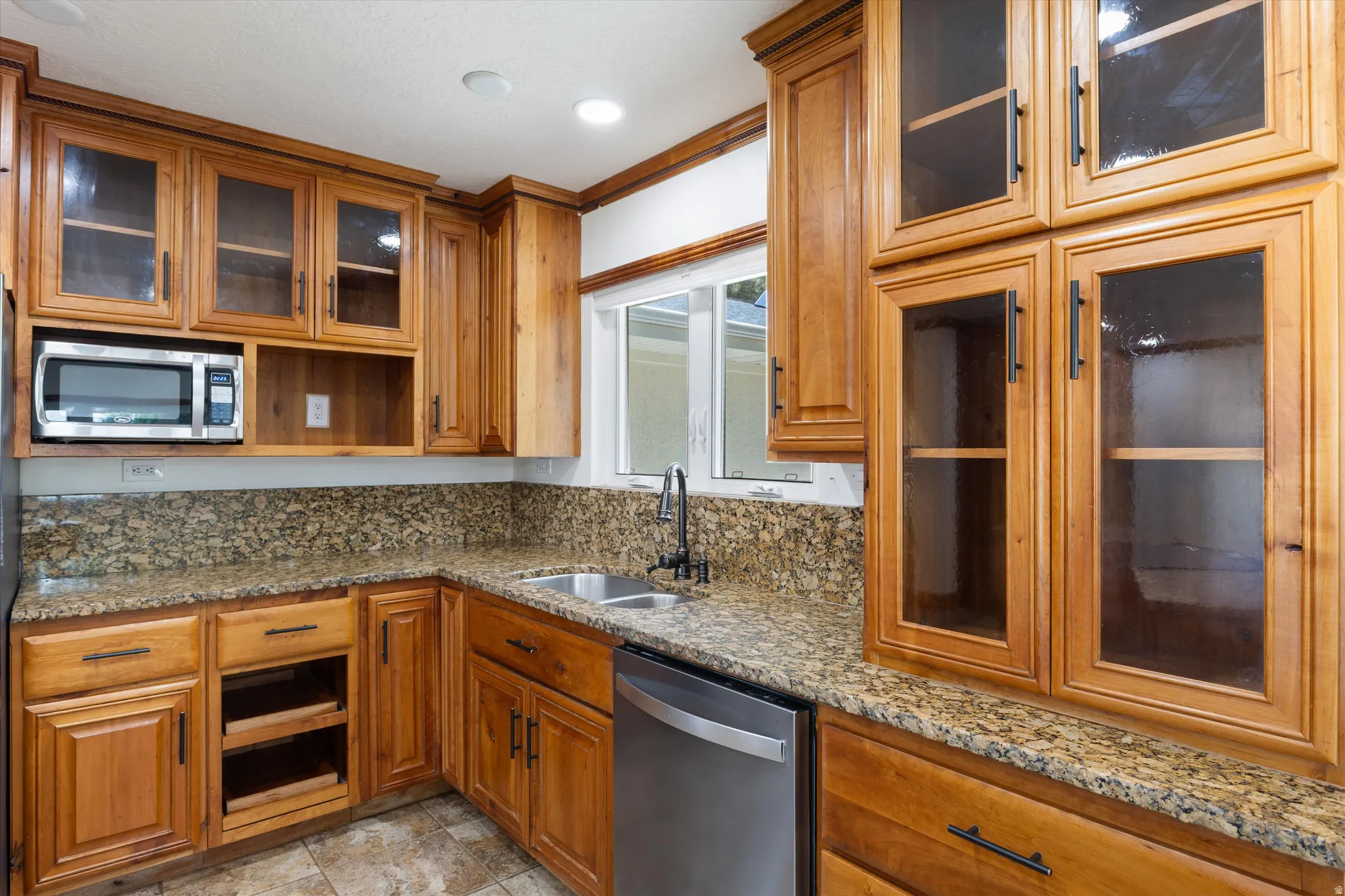 Light wood-finish cabinetry with glass-front uppers, granite-style countertops with backsplash, and a stainless steel dishwasher