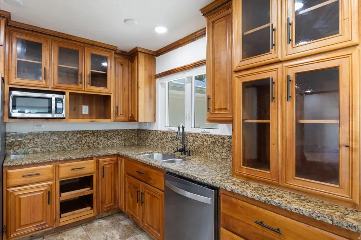 Light wood-finish cabinetry with glass-front uppers, granite-style countertops with backsplash, and a stainless steel dishwasher