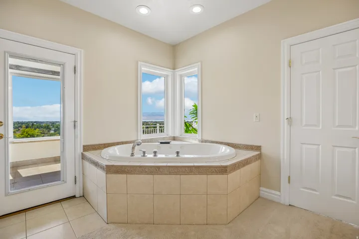 Corner jetted tub with tile surround, dual windows, and an exterior balcony access door