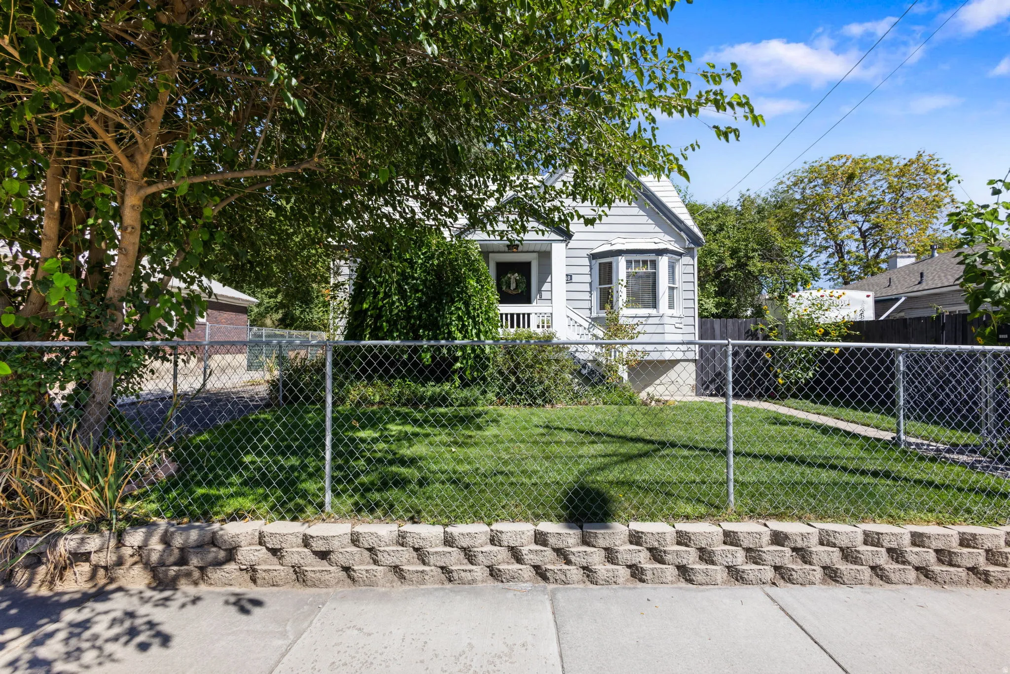 Siding exterior with a bay window, covered porch, chain link fence, retaining wall, and mature landscaping