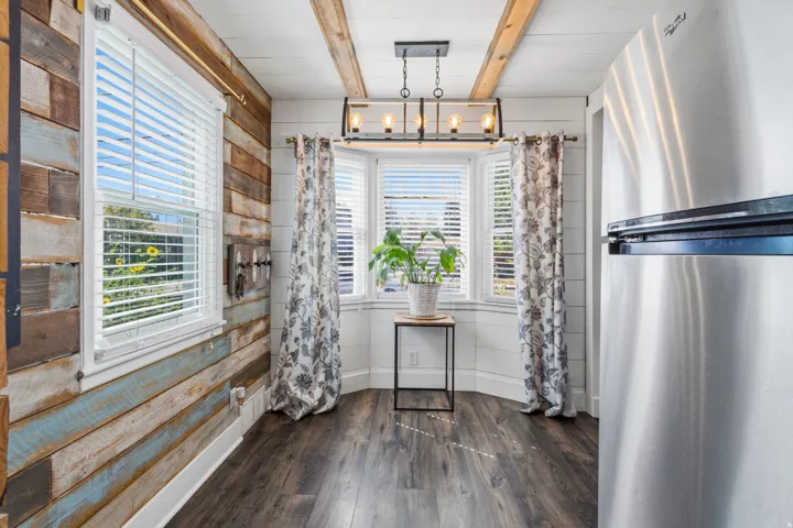Wood-finish flooring, accent wall with varied wood paneling, bay window, exposed ceiling beams, and a linear chandelier