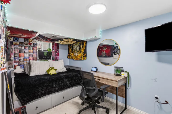 Built-in bed frame with storage drawers, light wood-finish desk with black metal frame, wall-mounted television, and a round wall mirror