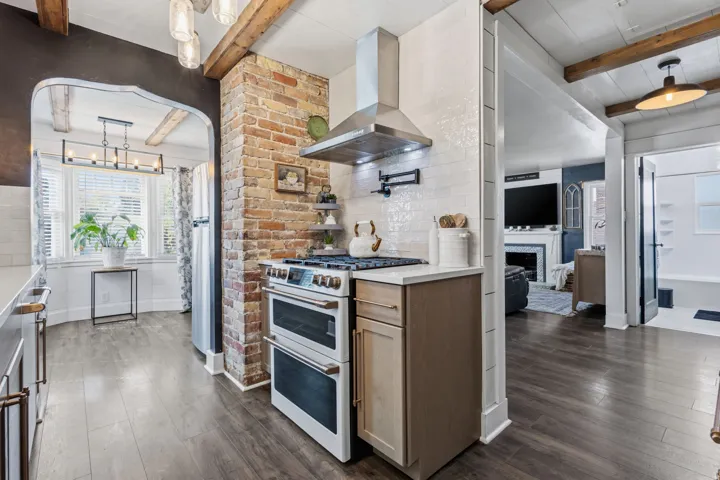 Kitchen featuring wood-finish flooring, exposed brick, white subway tile backsplash, stainless steel range with hood, and wood beam ceilings