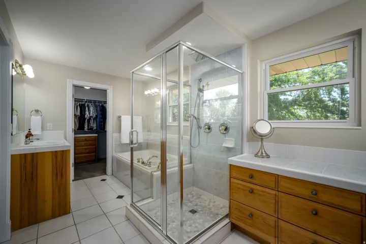 Walk-in shower with clear glass enclosure, a built-in bathtub, and dual wood-finish vanity cabinets with integrated sinks and a walk in closet on each side