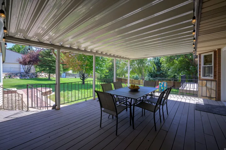 Covered outdoor deck with a metal patio roof, composite decking, and wrought iron railings, overlooking a grassy yard with mature trees