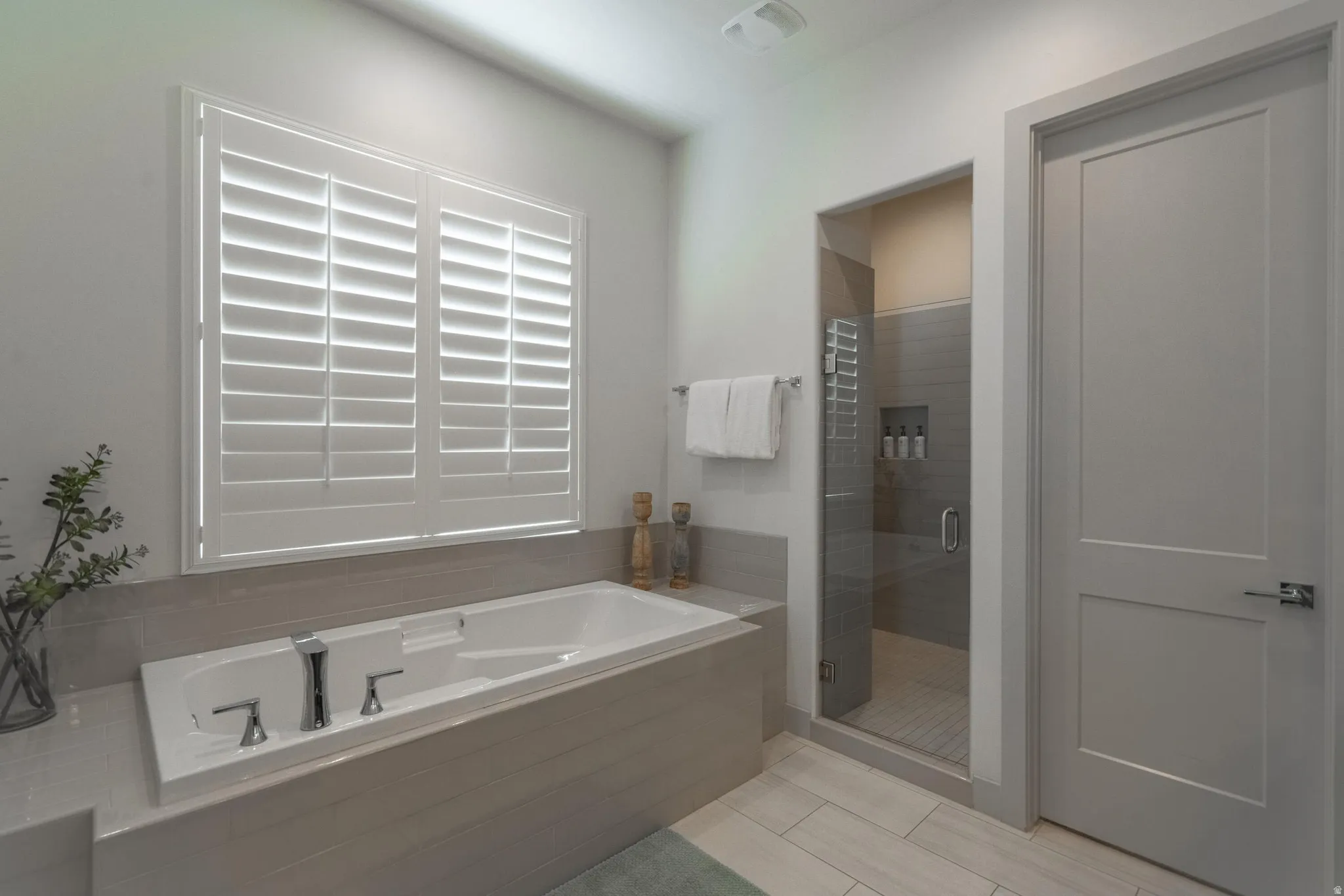 Main Floor Primary Ensuite Built-in soaking tub with chrome fixtures, plantation shutters, and a glass-enclosed shower with tiled walls and a recessed niche
