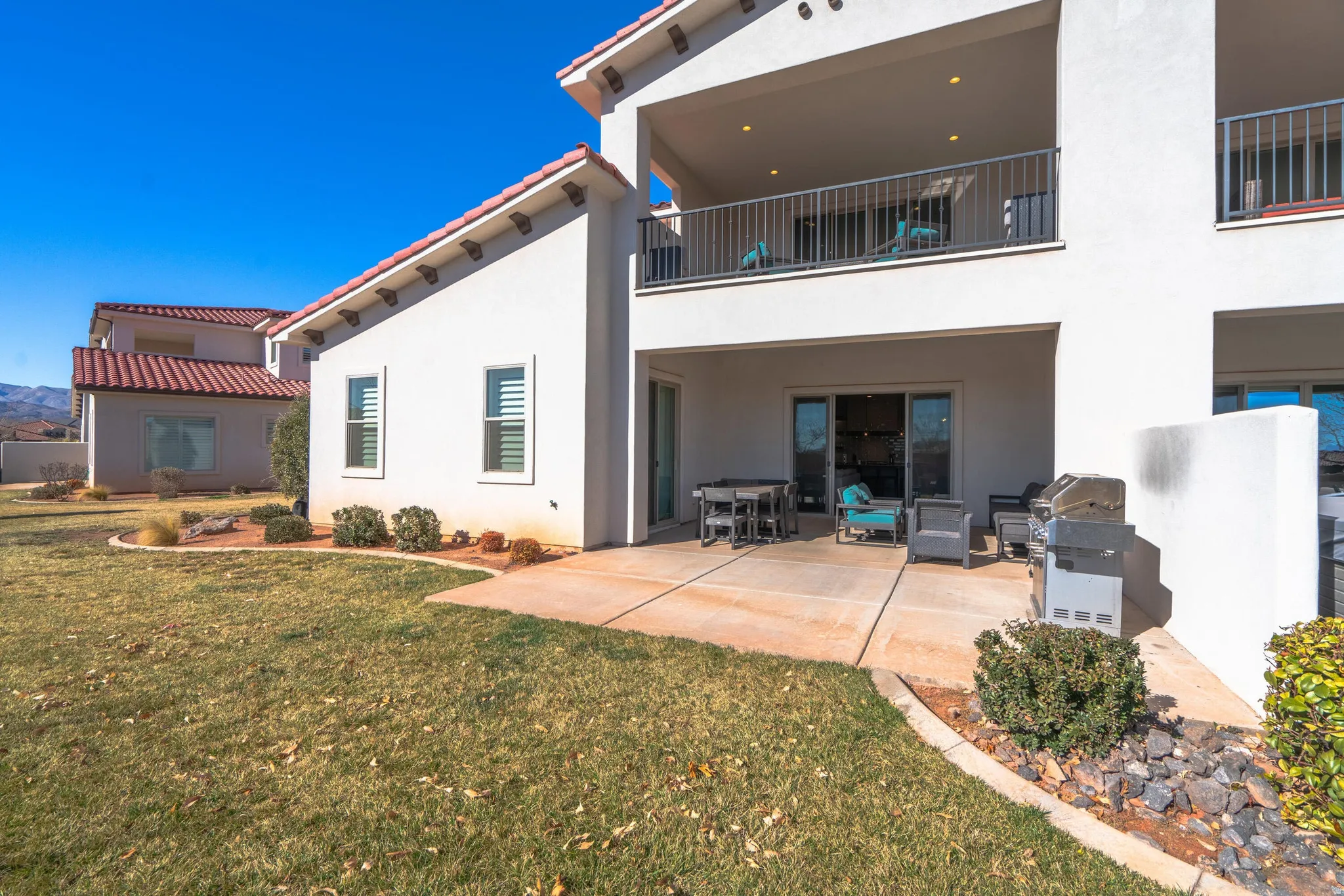 Expansive outdoor patio with concrete paving, sliding glass door access, an upper-level balcony with railings, and landscaping with shrubs and natural rock accents