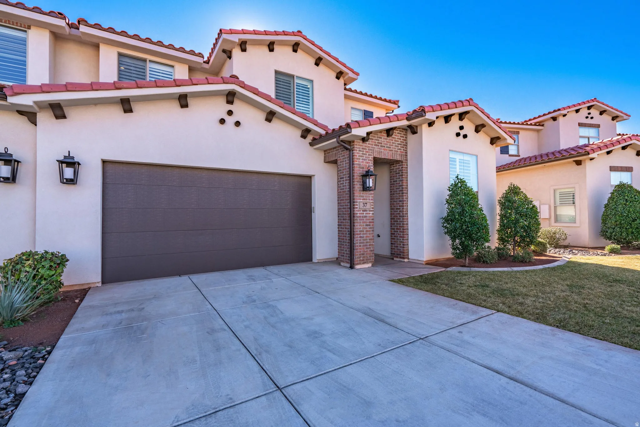 Stucco exterior with a tile roof, an integrated garage, brick entry accents, and mature landscaping