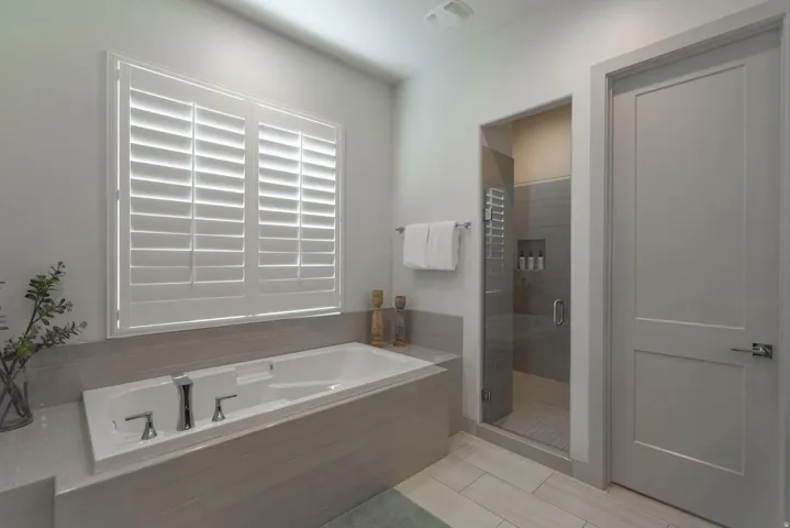 Main Floor Primary Ensuite Built-in soaking tub with chrome fixtures, plantation shutters, and a glass-enclosed shower with tiled walls and a recessed niche