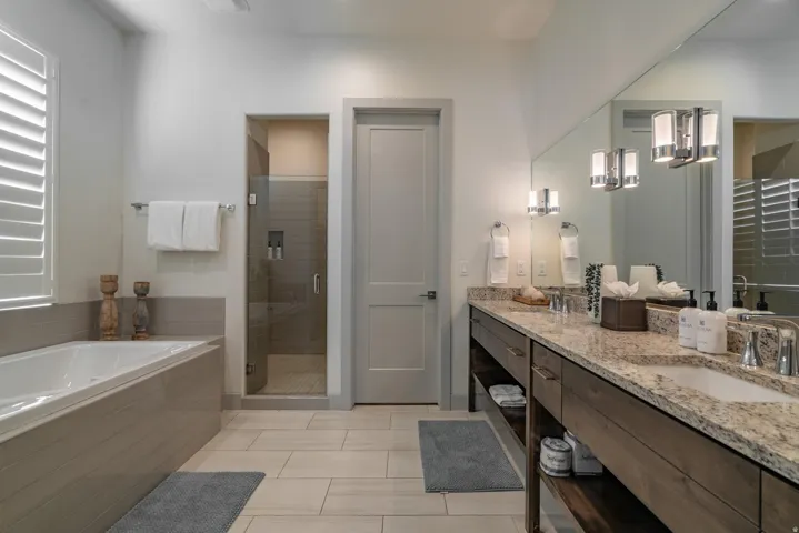 Main Floor Primary Ensuite Dual vanity with granite countertop, integrated soaking tub, separate glass-enclosed shower, wood-finish cabinetry, and neutral tile flooring